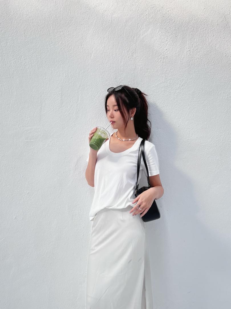 Huan Min in a white outfit holding a matcha drink against a sunlit wall.