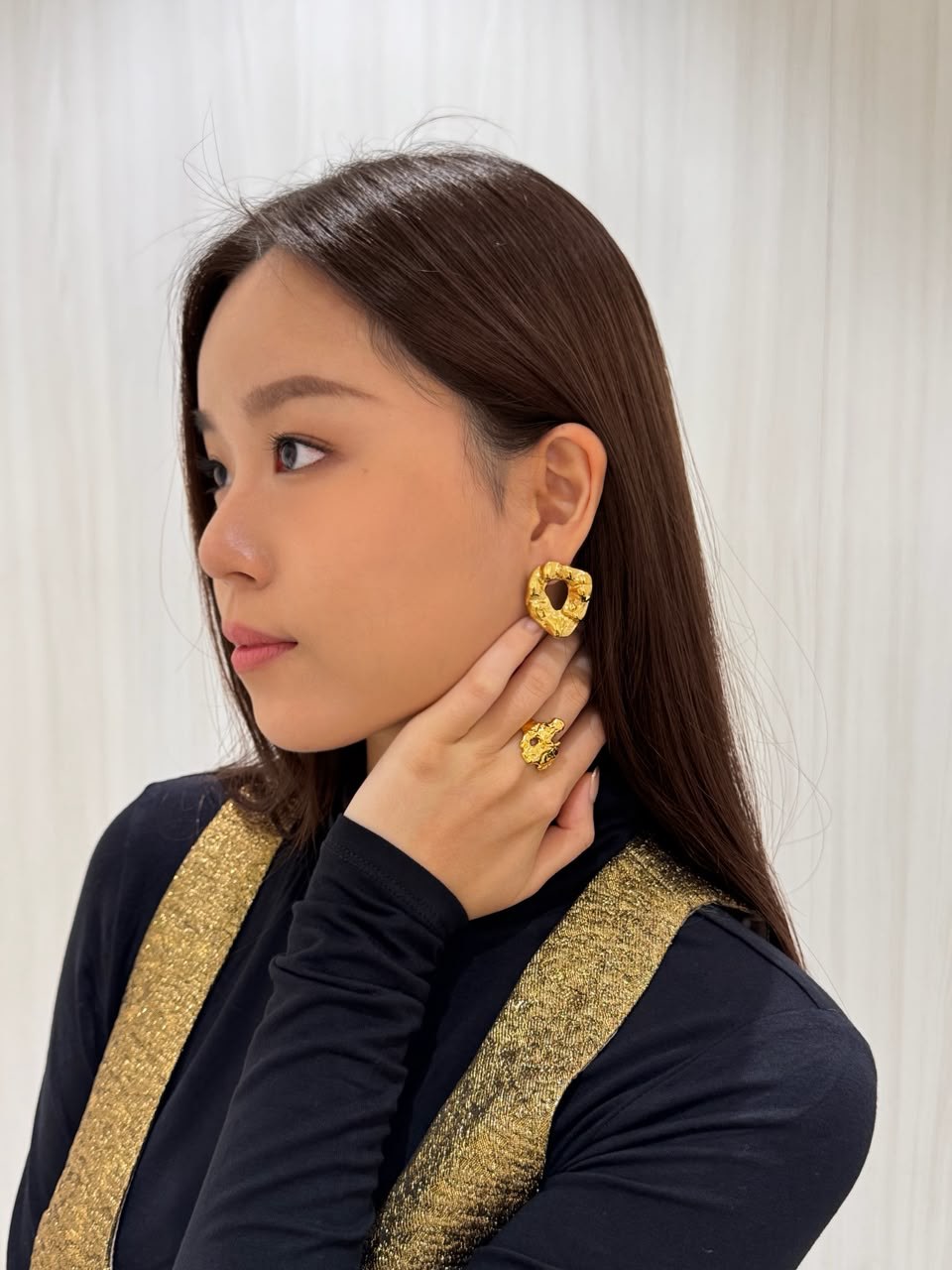 Huan Min in a black-and-gold outfit, presenting gold jewellery with her hand beside her face.